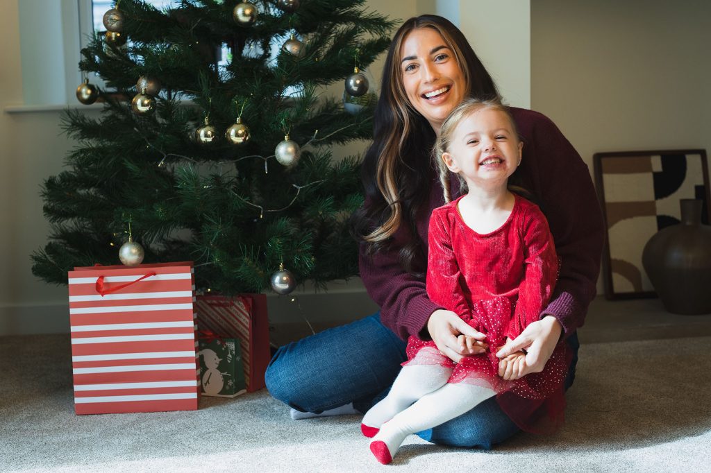 A young girl in a red top sat on her mothers lap by a Christmas tree