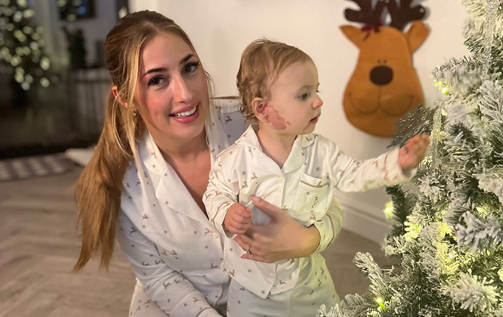 A young girl being held by her mother near a Christmas tree. Both are wearing matching white pyjamas.